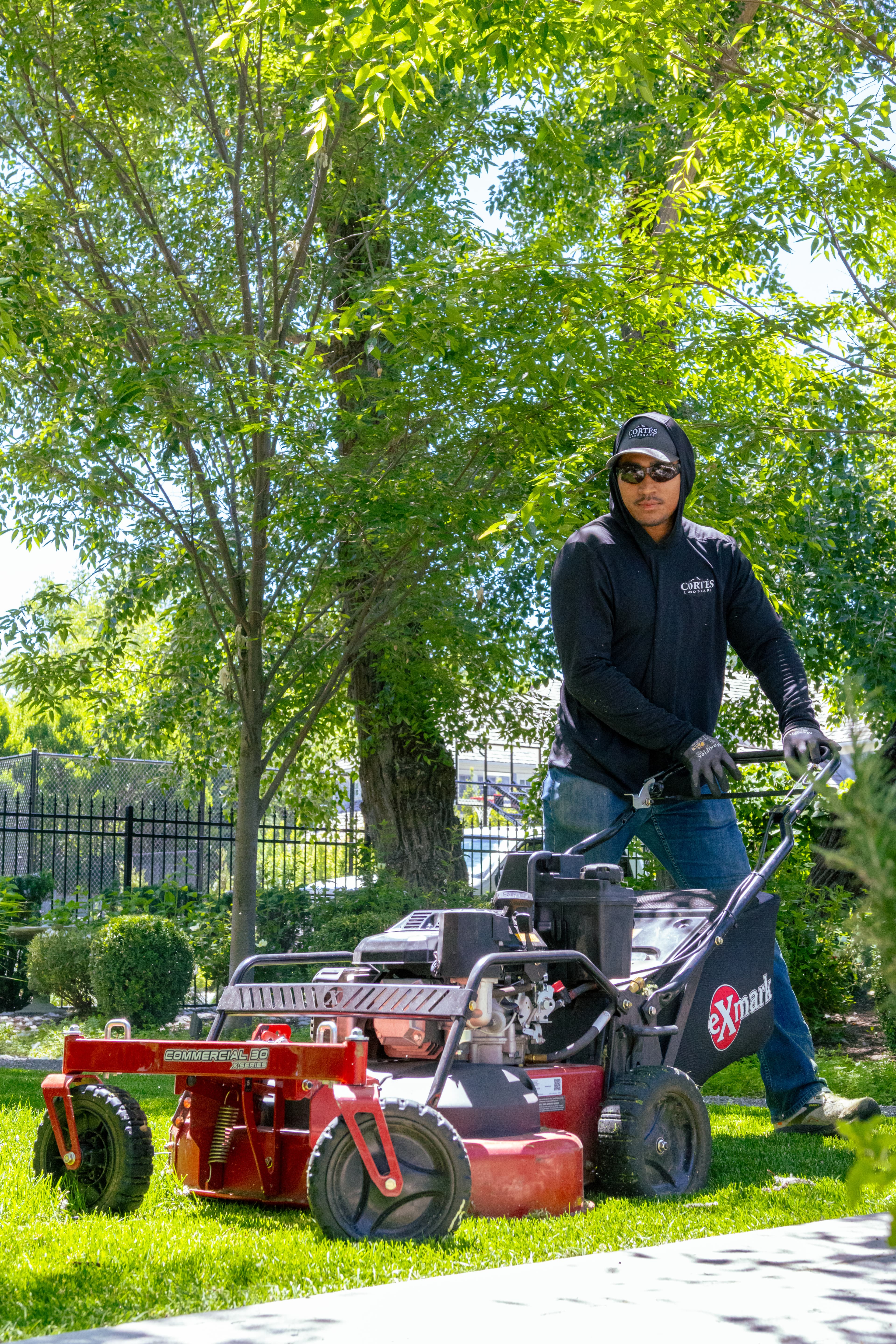 Man in black hoodie and sunglasses operating a red commercial lawn mower on green grass.