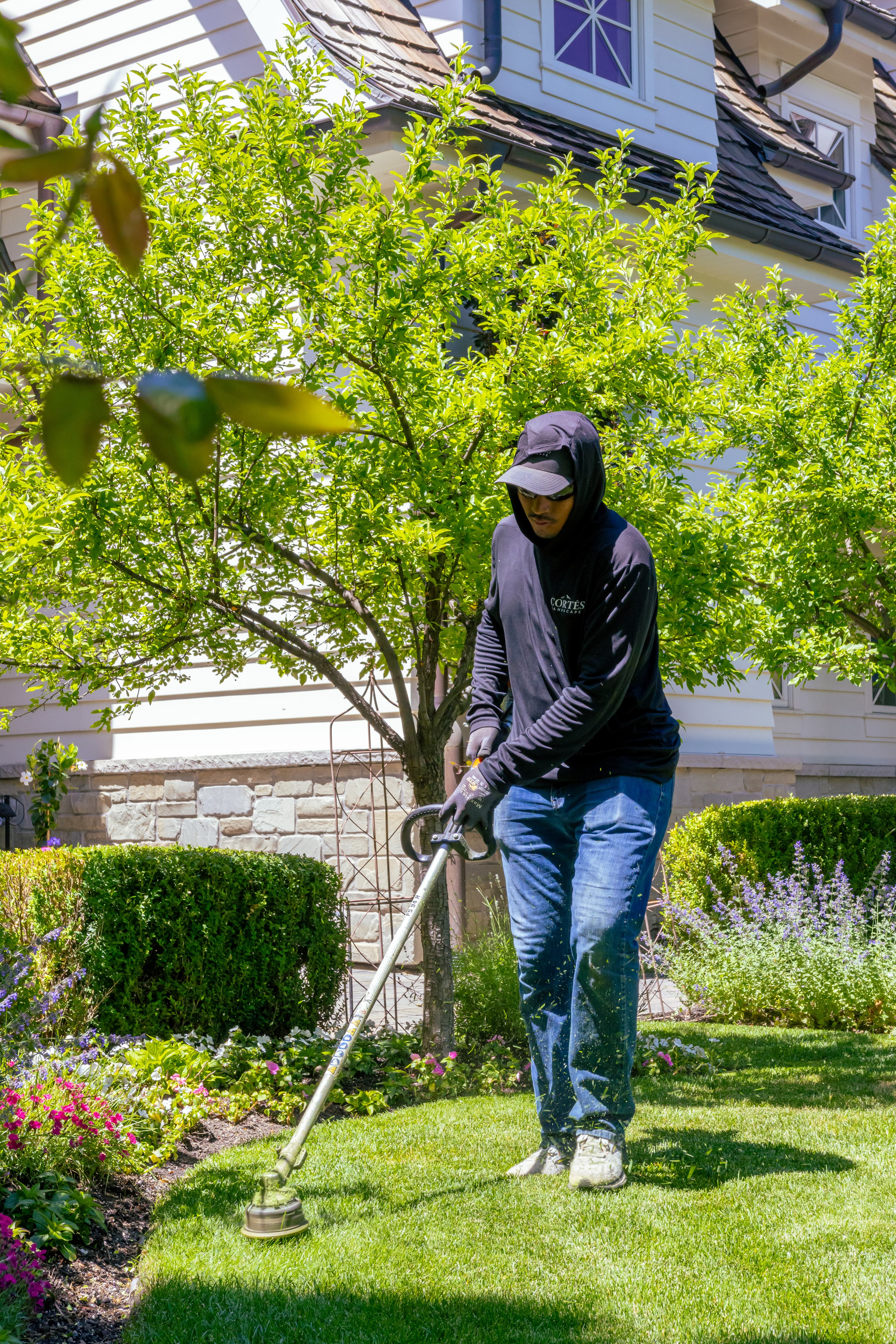 Landscaper in a black hoodie uses a string trimmer to edge a sunny green lawn.