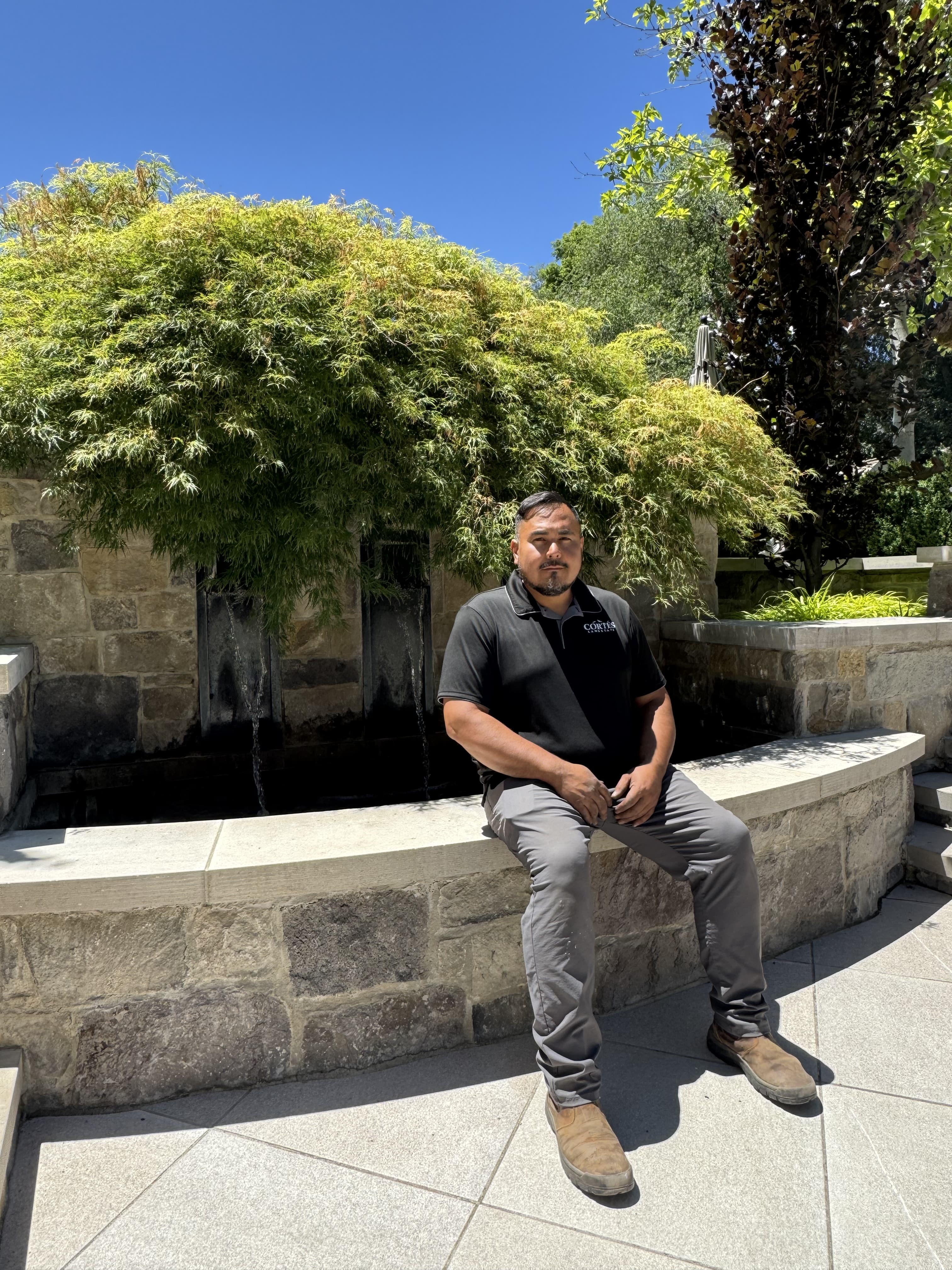 Man in black polo sits on a stone ledge before a waterfall and weeping tree.
