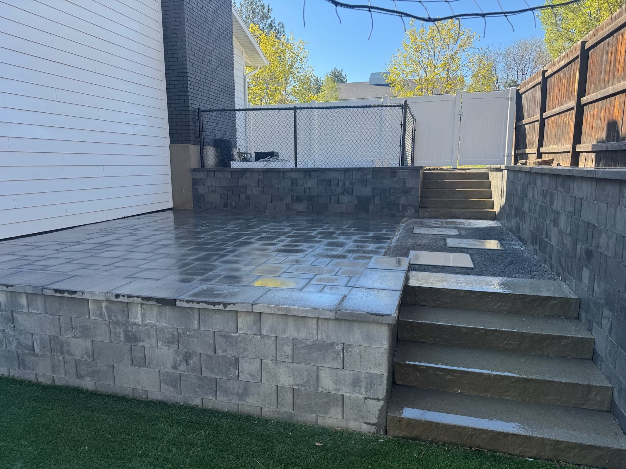 Modern tiered gray stone patio with steps and retaining walls in a fenced backyard.